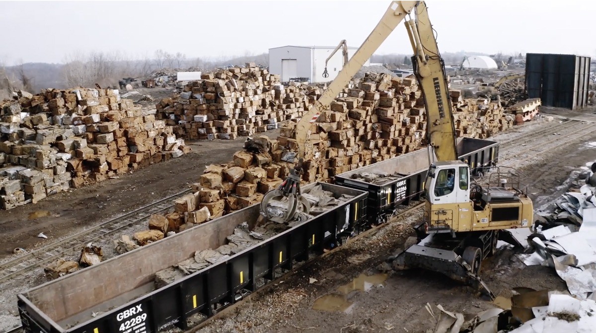 A yellow crane in an industrial yard loading a GBRX railcar